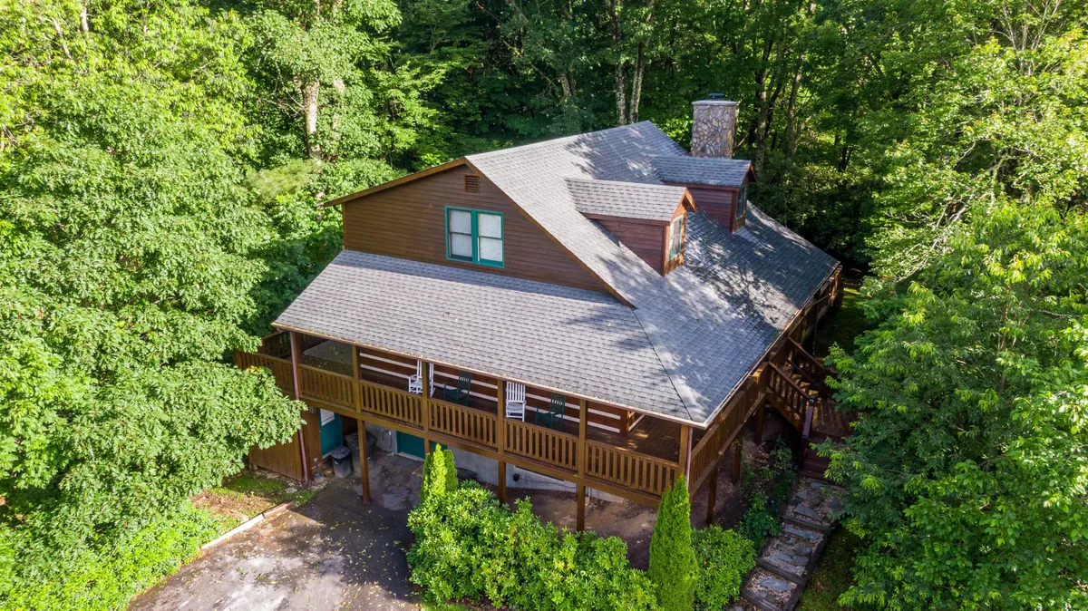 This image features a welcoming wooden porch with a prominent stone chimney and is surrounded by lush trees, inviting guests to step outside and experience the natural environment. The spacious porch is ideal for gathering, relaxing, and soaking in the serenity of the Blue Ridge Mountains.