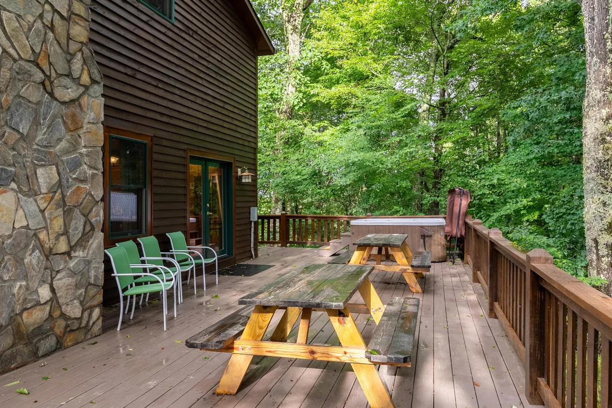 This image provides a wide view of the spacious wooden deck featuring picnic tables, a hot tub, and green chairs set against a backdrop of trees and a stone wall. The space appears ready for a family meal, group gathering, or a relaxing evening in the hot tub, all while immersed in a natural mountain setting.