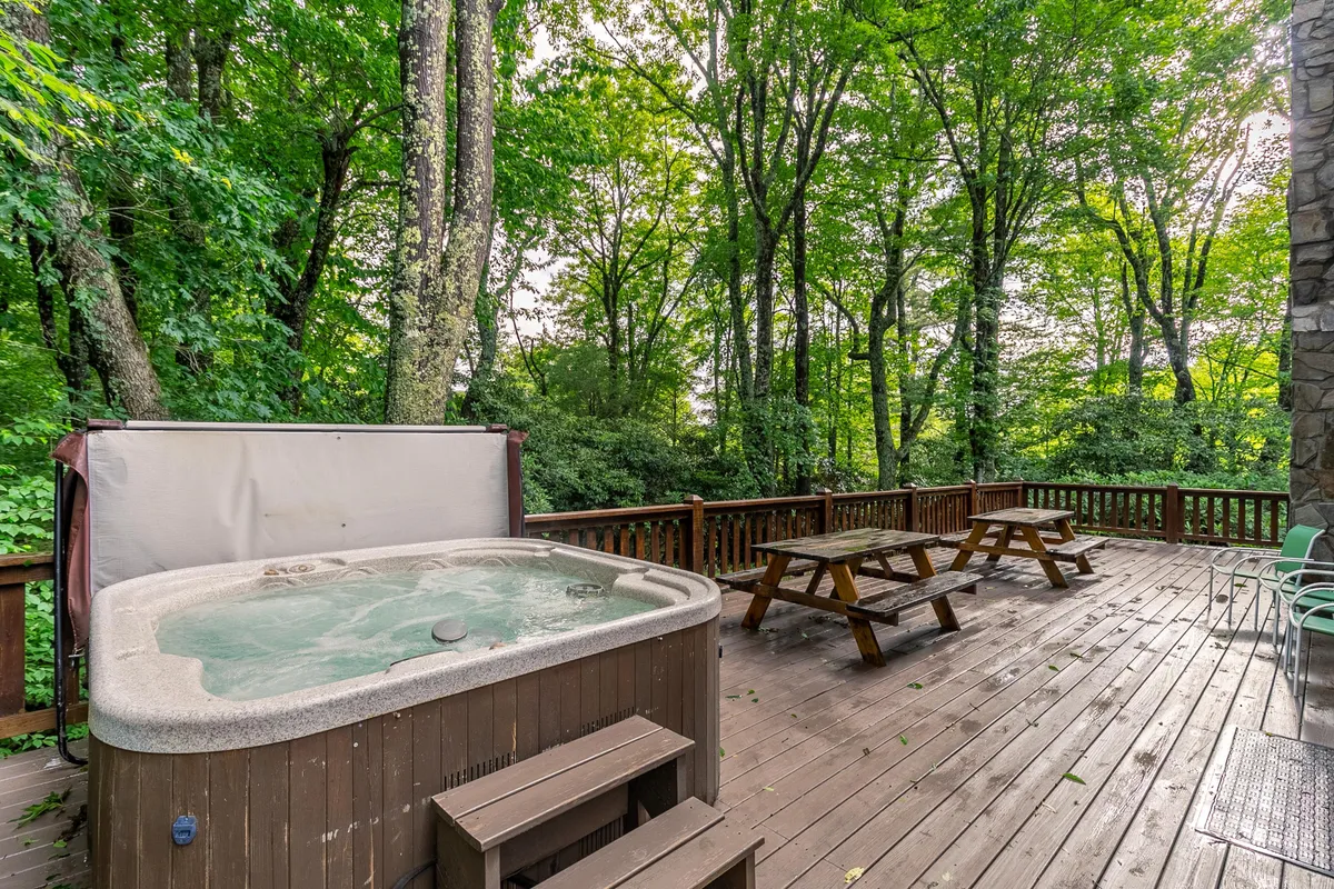 The image highlights the inviting outdoor hot tub nestled on the deck, with picnic tables nearby and surrounded by lush trees. The setting is quiet and peaceful, perfect for socializing, relaxing under the stars, or enjoying coffee in the morning mountain air.