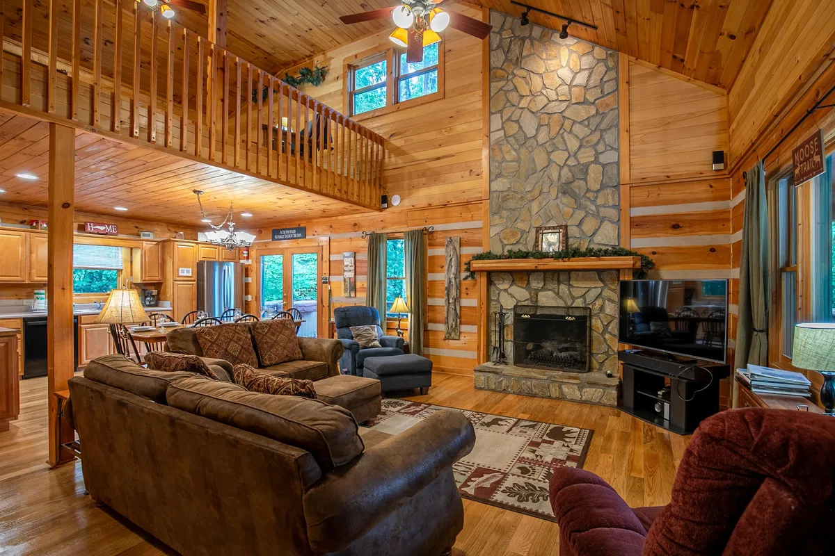 A cozy interior living space with a stone fireplace, comfortable seating area, exposed wooden beams, and large windows letting in natural light. The space exudes warmth and is perfect for relaxing, working by the fire, or enjoying family time.