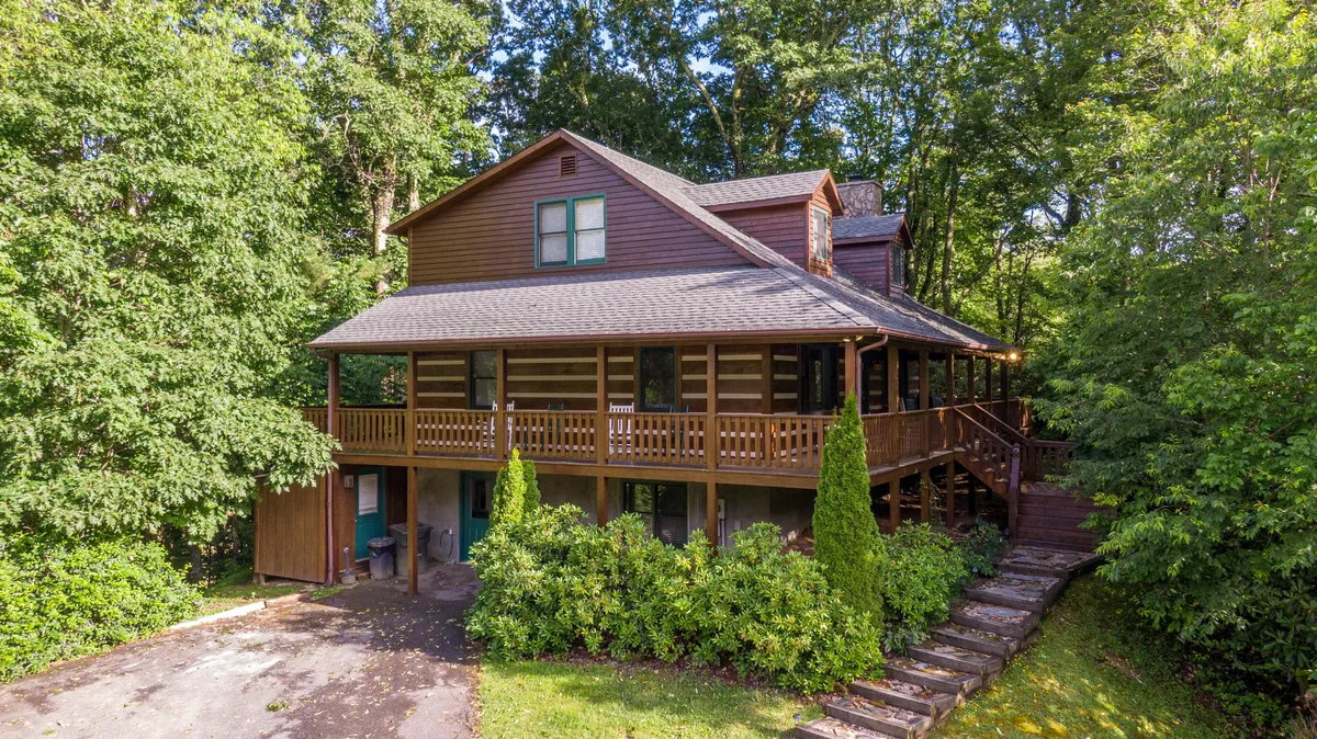 A classic log cabin nestled in the lush greenery of Boone, North Carolina; the hero image features a charming wrap-around porch, rustic stone pathway, and inviting outdoor spaces perfect for family gatherings or relaxing in nature.