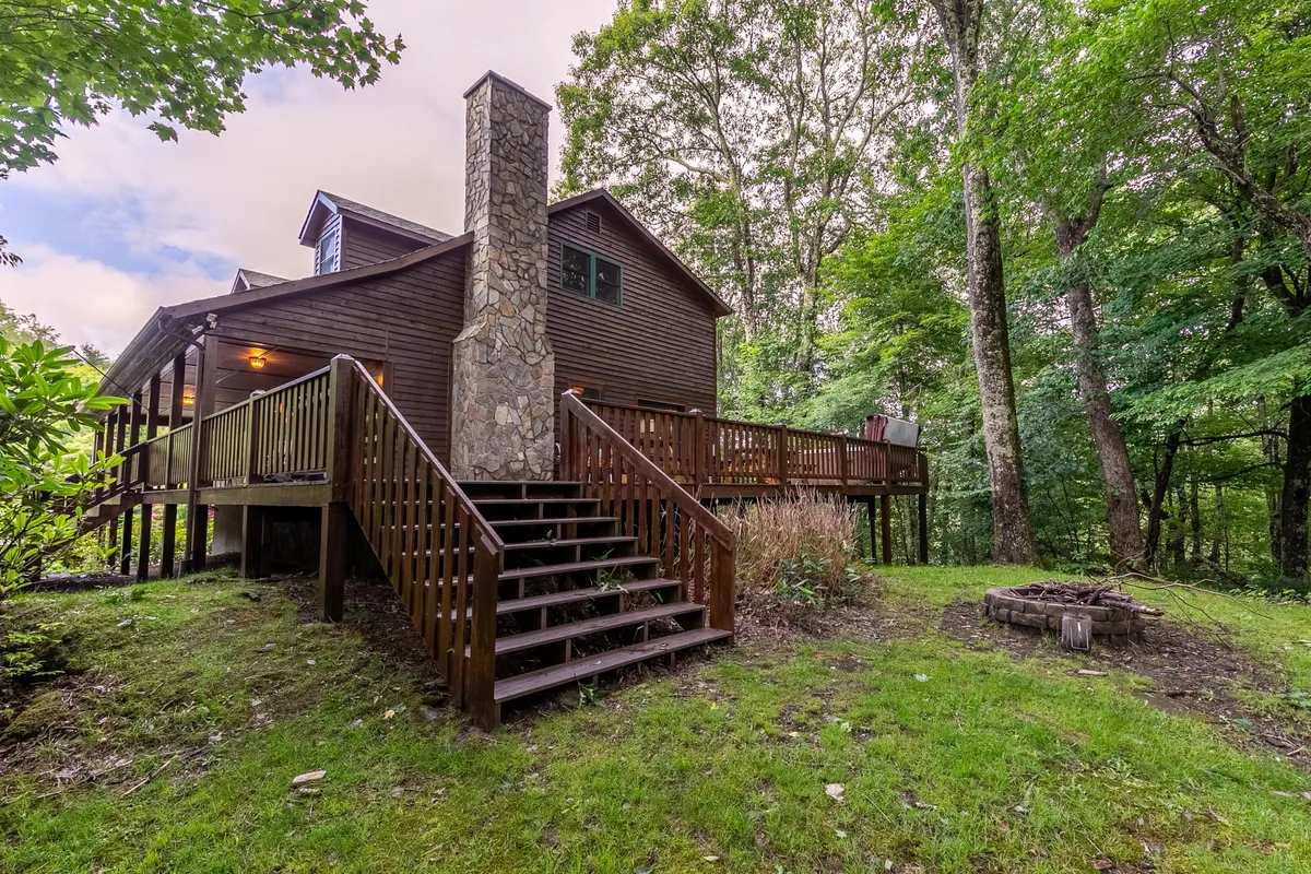 This image features the cabin's exterior, highlighting a spacious deck with wooden stairs, a stone chimney, a fire pit area, and lush wooded surroundings. The outdoor seating and natural setting emphasize the connection to nature and the inviting space for gatherings after a day of adventure.