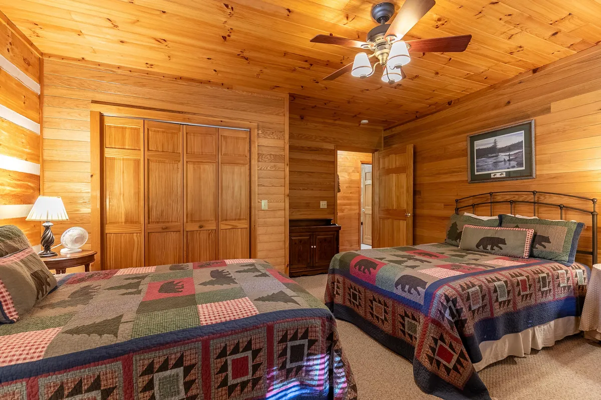 A bedroom with bedding featuring playful bear patterns, surrounded by wooden paneling. The room is brightened by natural sunlight, and includes a ceiling fan for added comfort, evoking a rustic, inviting feel.