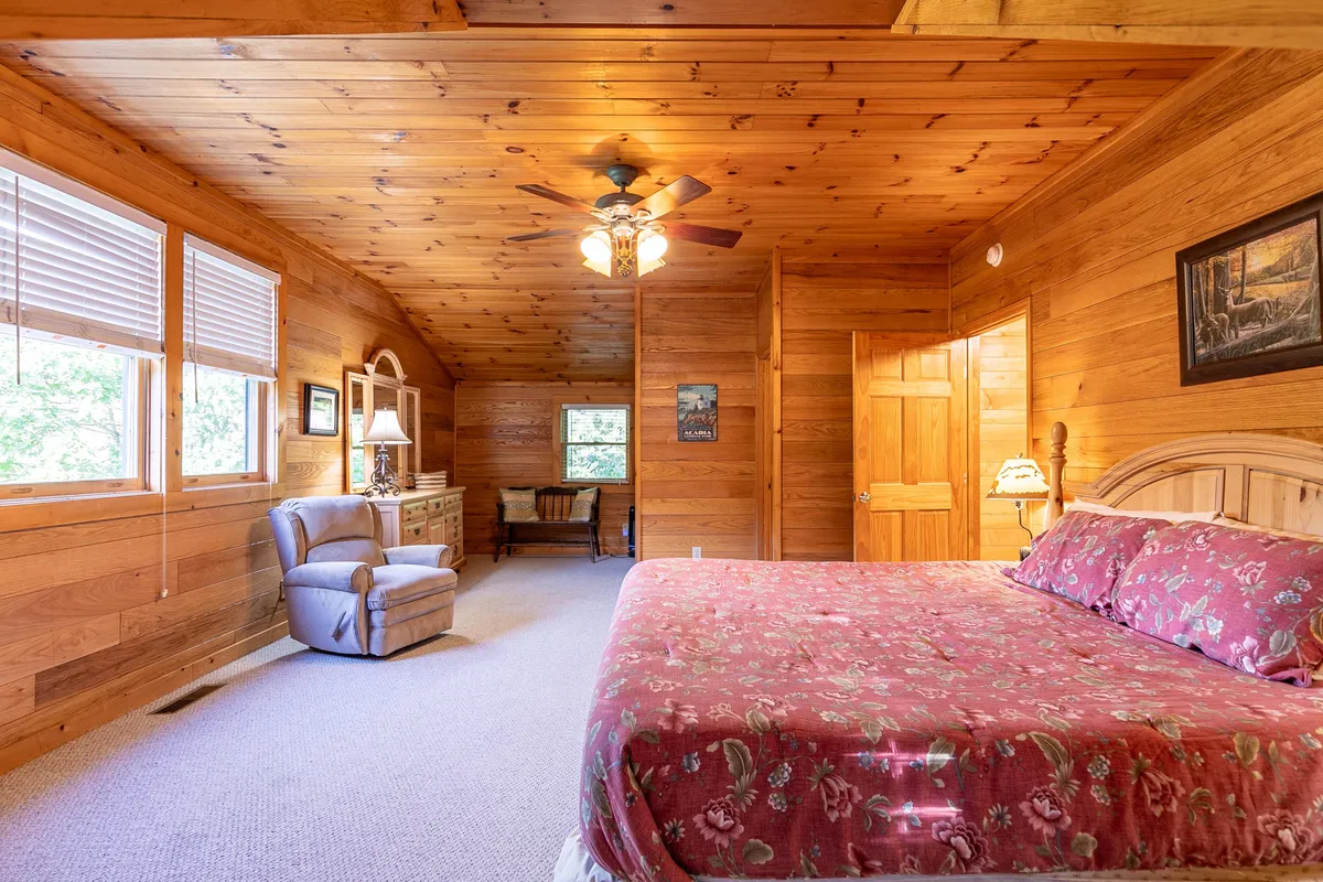 A restful bedroom with floral bedding, wooden ceiling and walls, a comfortable armchair, and a dresser with a lamp. The ambiance is calm and homey, inviting moments of relaxation and peaceful mornings in the mountains.