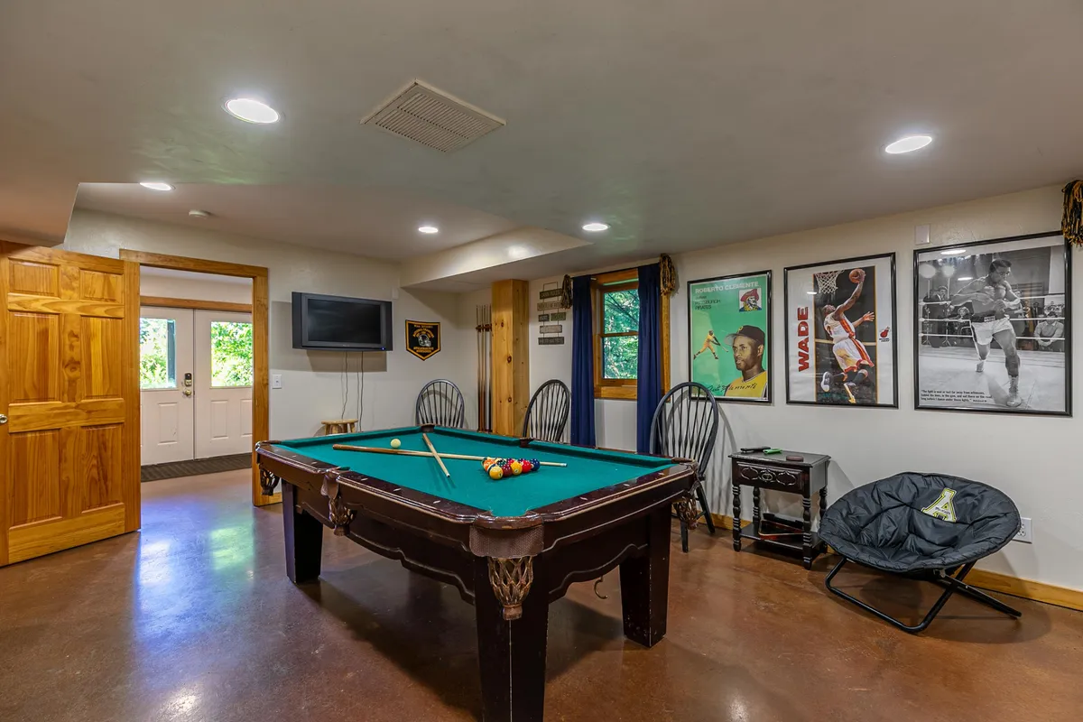 A vibrant lower-level game room featuring a pool table at the center, modern wall art, and a comfortable seating area. The space is set up for friendly competitions and casual gatherings, with a relaxed and inviting ambiance perfect for socializing or enjoying a game night.