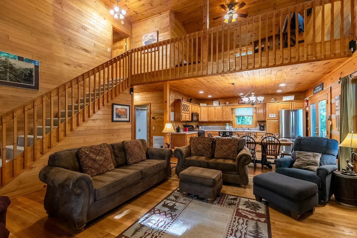 A wide view of the open-plan main living area, featuring a central staircase, plush sofas, a spacious dining area, and a modern kitchen in the background. The space is bathed in warm light, emphasizing a welcoming and communal atmosphere ideal for gatherings and entertaining.