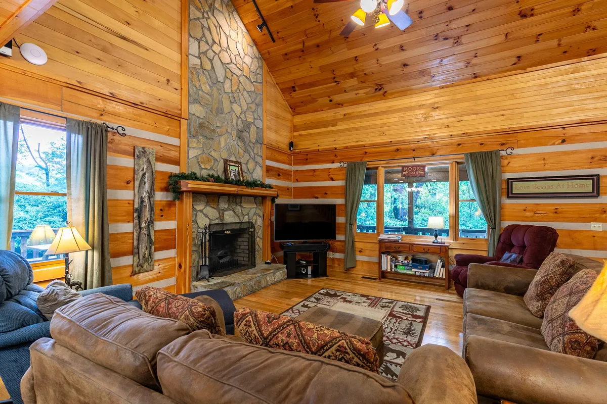 A cozy living room anchored by a towering stone fireplace, large windows offering plenty of natural light, comfortable seating, and a decorative rug. The setup encourages relaxation, socializing, and enjoying entertainment together.